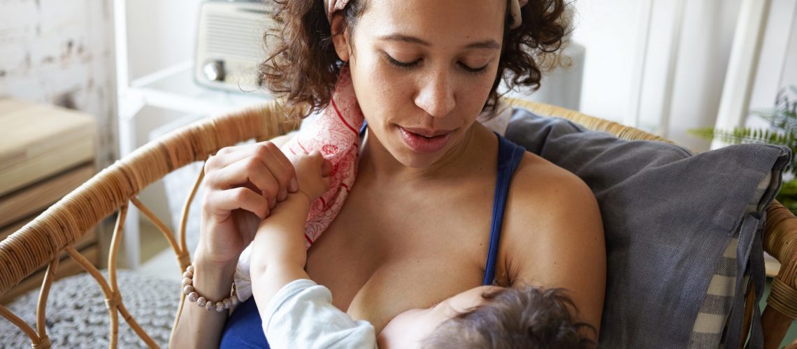 Infancy, motherhood, nutrition, lactation and breastfeeding concept. Indoor portrait of tender attractive young mom nursing her baby boy, giving him all love and affection, sitting in bedroom